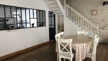 Un coin repas avec une table dressée pour quatre personnes, des chaises blanches, un plancher en bois, un escalier avec des rampes blanches et un mur en pierre.