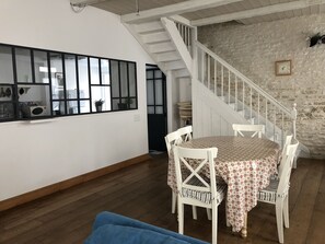 A dining area with a table set for four, white chairs, a wooden floor, a staircase with white railings, and a stone wall.