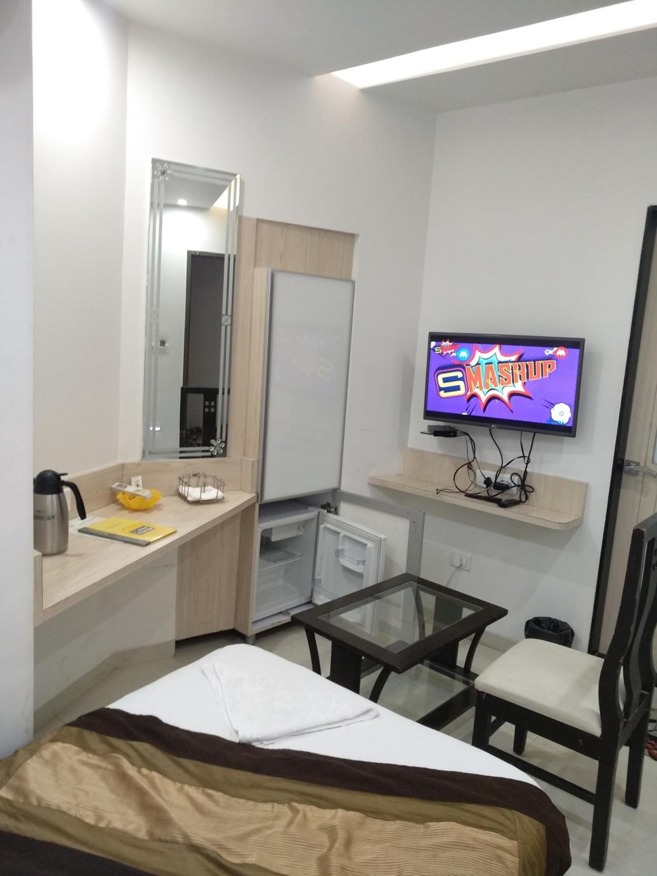A hotel room with a bed, a small table, a chair, a TV, a mirror, a mini-fridge, and a shelf with a kettle and a yellow object.