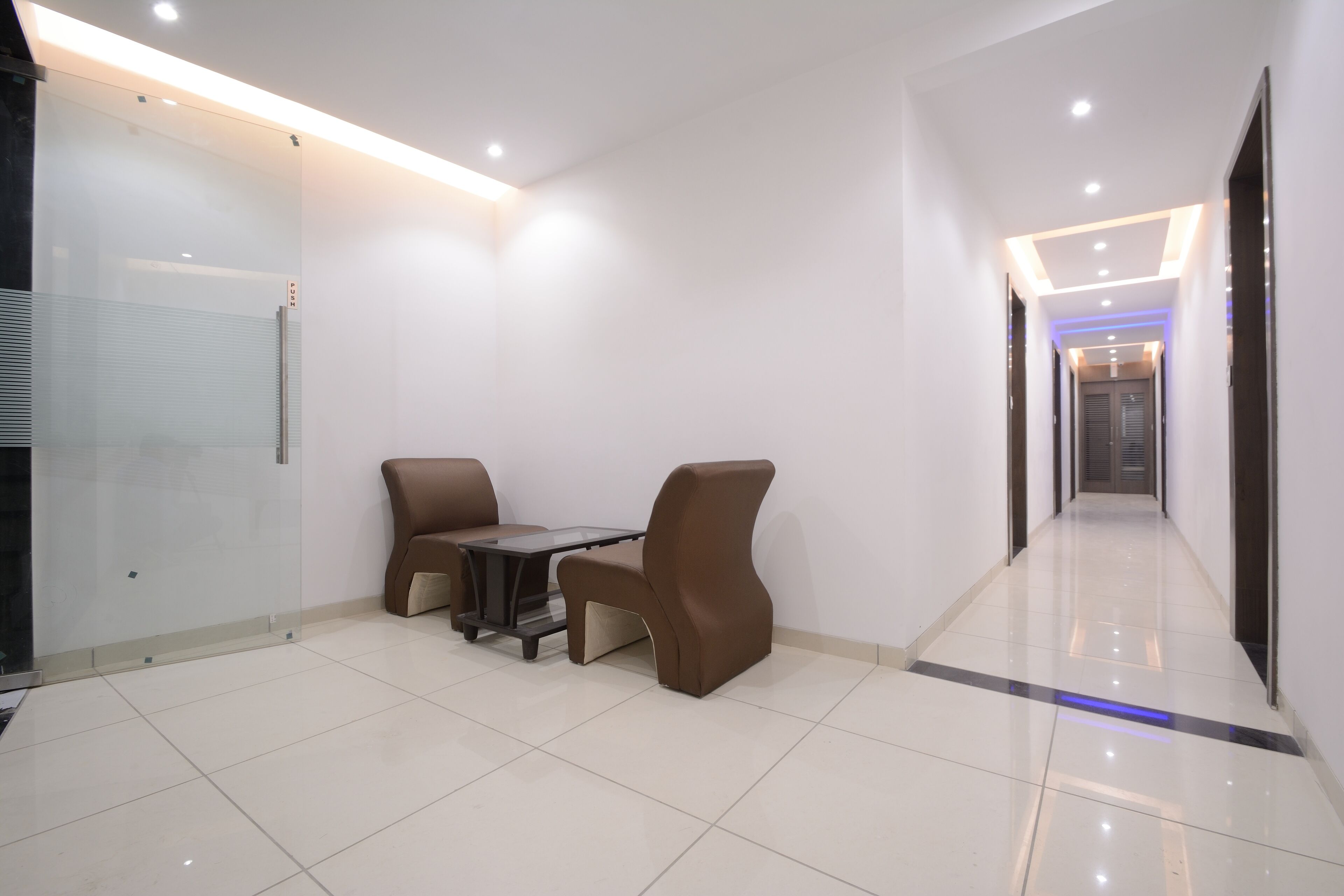 A modern hotel lobby with a glass partition, a seating area with a table and chairs, and a long corridor with recessed lighting.