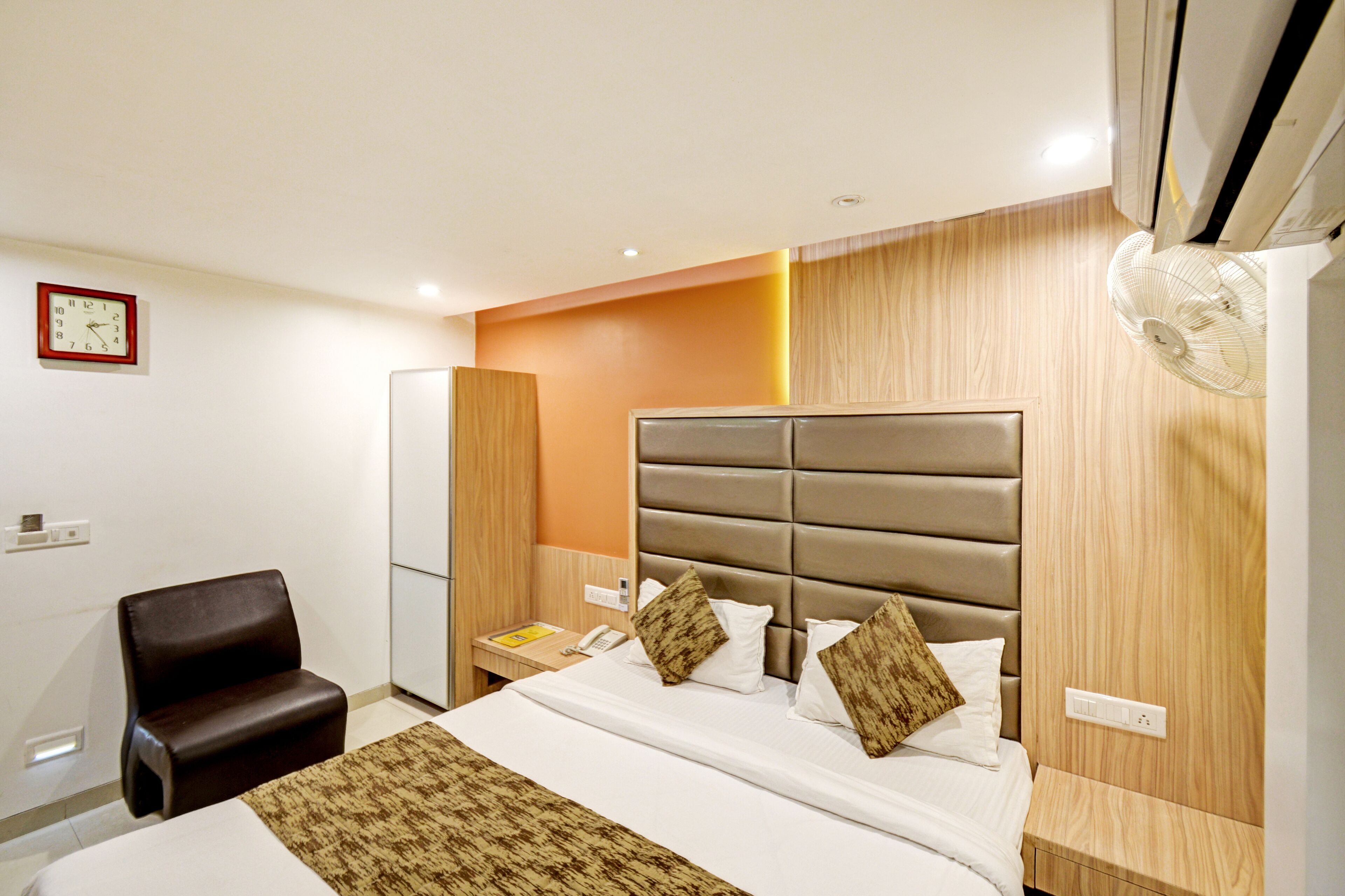 A hotel room with a bed, a chair, a bedside table, a clock, and an air conditioner.