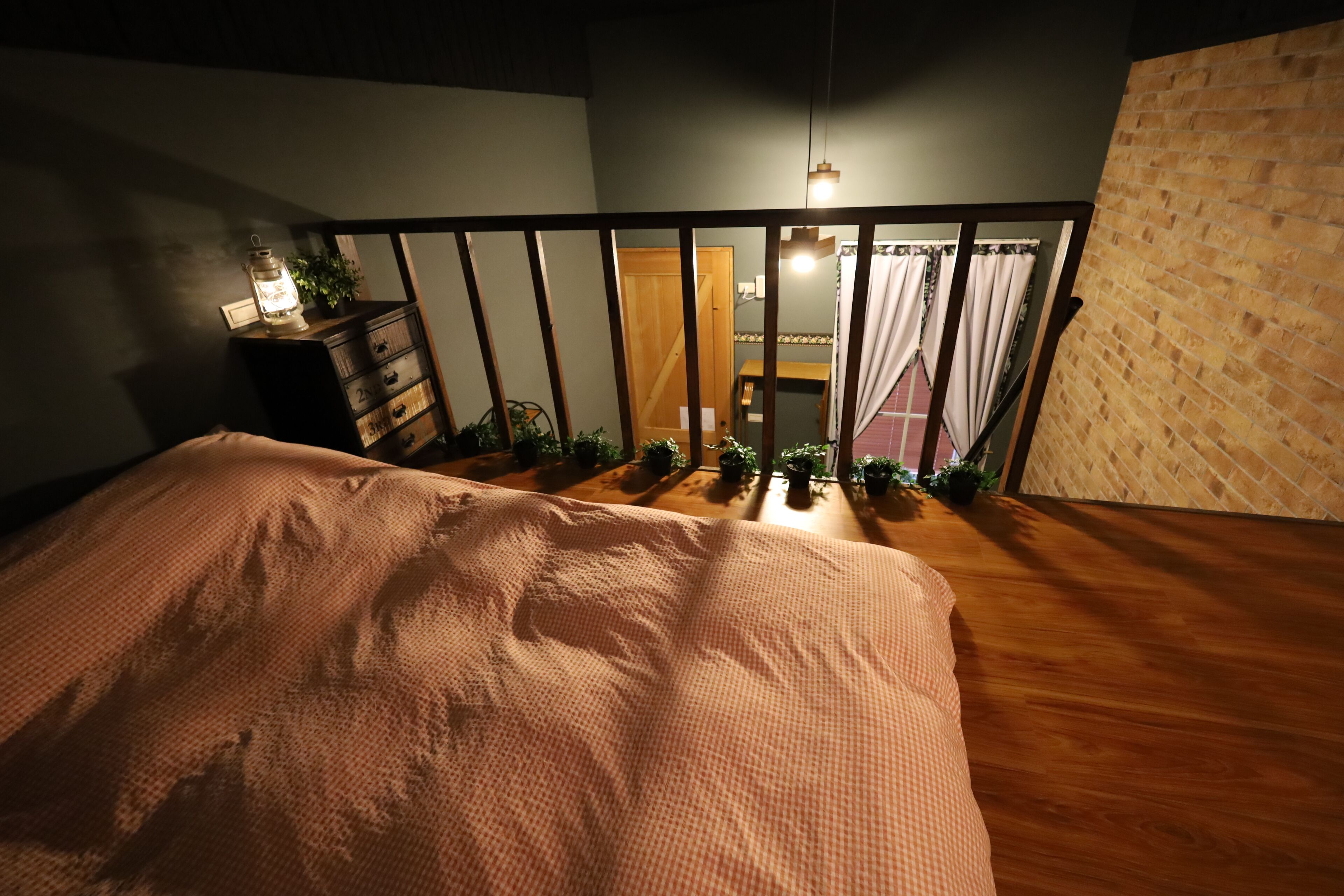 A bedroom with a wooden floor, a bed with a patterned bedspread, a nightstand with a lantern, and a view of a balcony with plants and a door.
