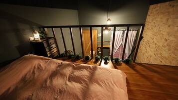 A bedroom with a wooden floor, a bed with a patterned bedspread, a nightstand with a lantern, and a view of a balcony with plants and a door.