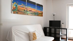 En vit soffa med en kudde, ett svart TV-bord och en färgstark, abstrakt tavla på väggen.