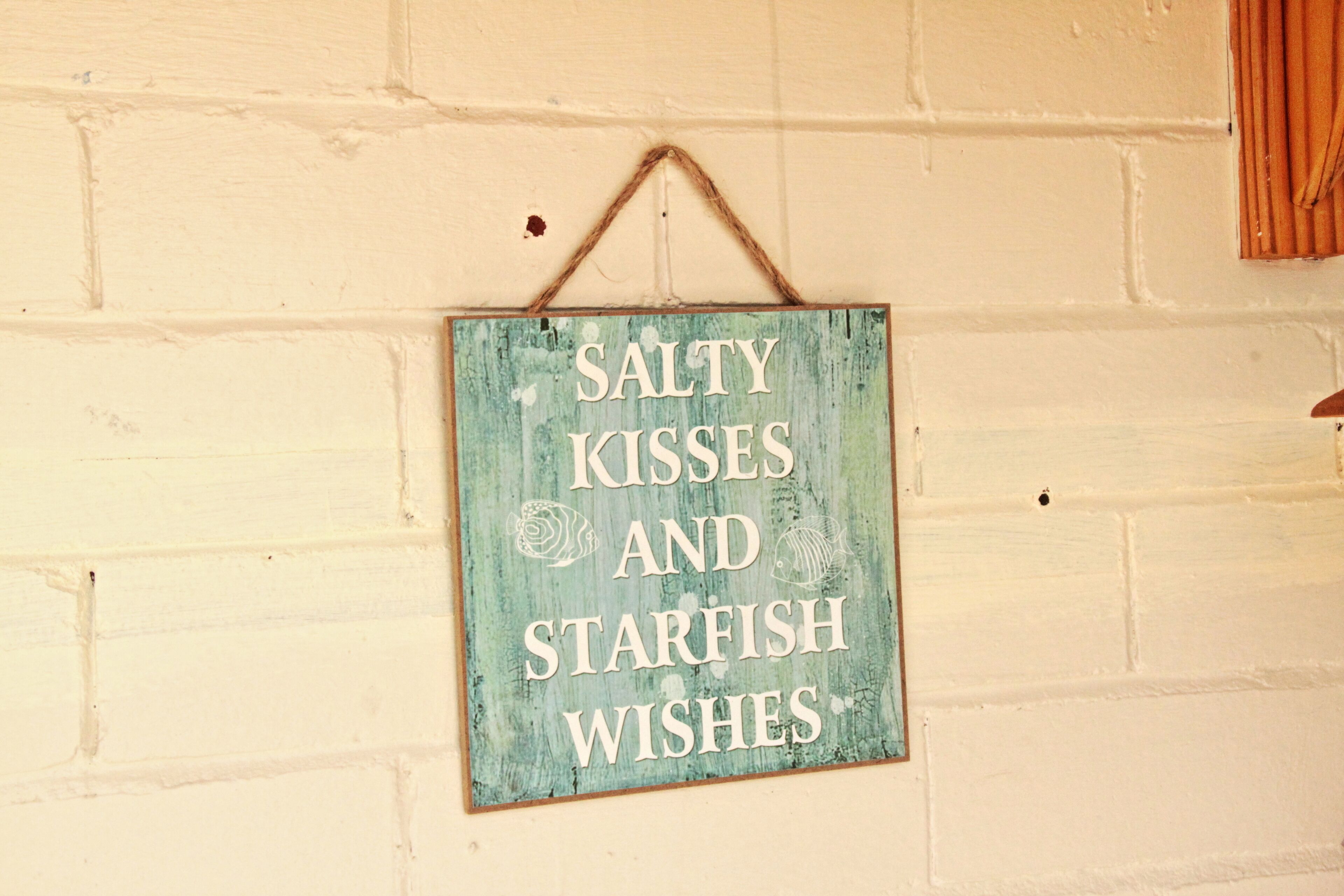 A rustic sign with the text "SALTY KISSES AND STARFISH WISHES" on a white brick wall.