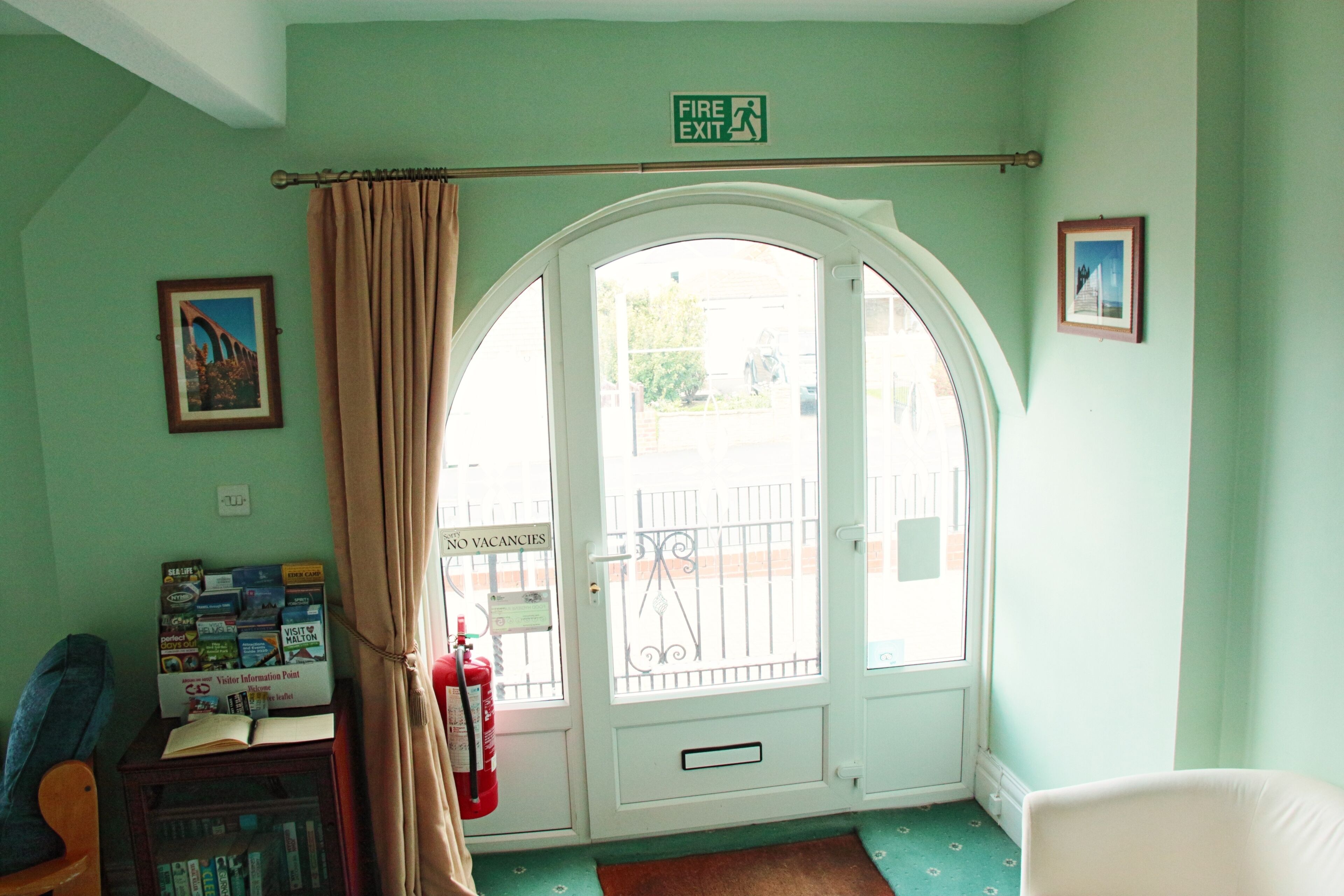 A room with a large arched window, a fire exit sign, a fire extinguisher, a chair, a small table with books, and framed pictures on the wall.