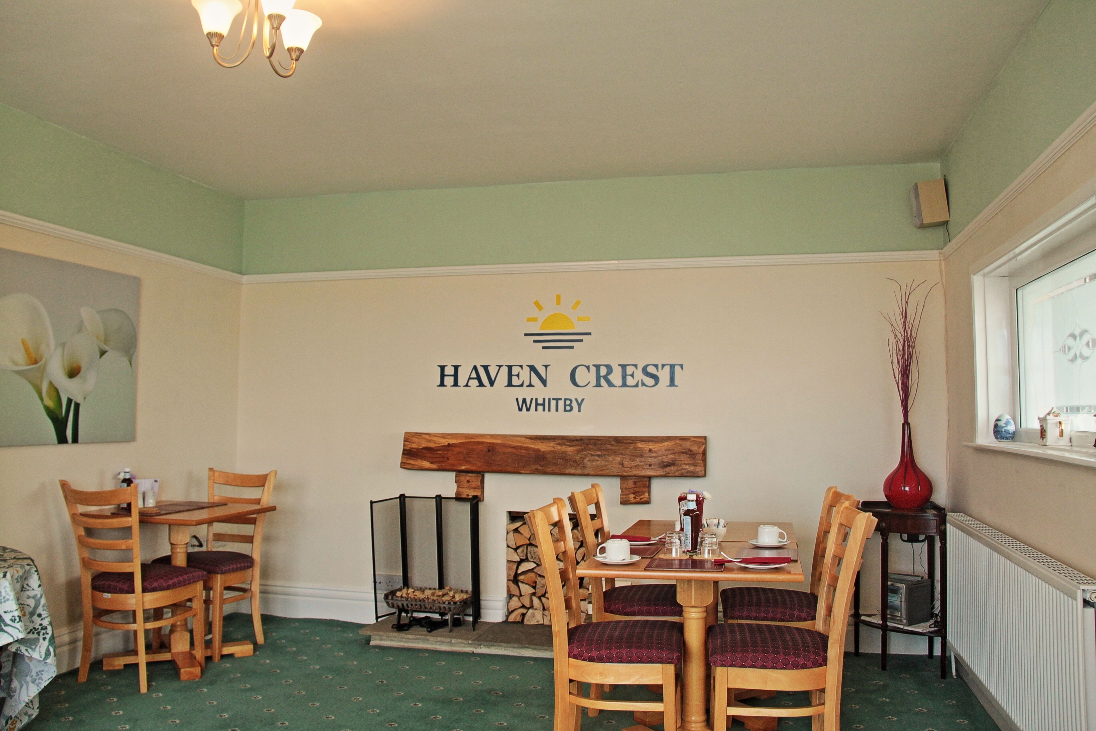 Uma área de refeições com mesa e cadeiras de madeira, lareira e um letreiro fixado na parede com a inscrição "HAVEN CREST WHITBY".