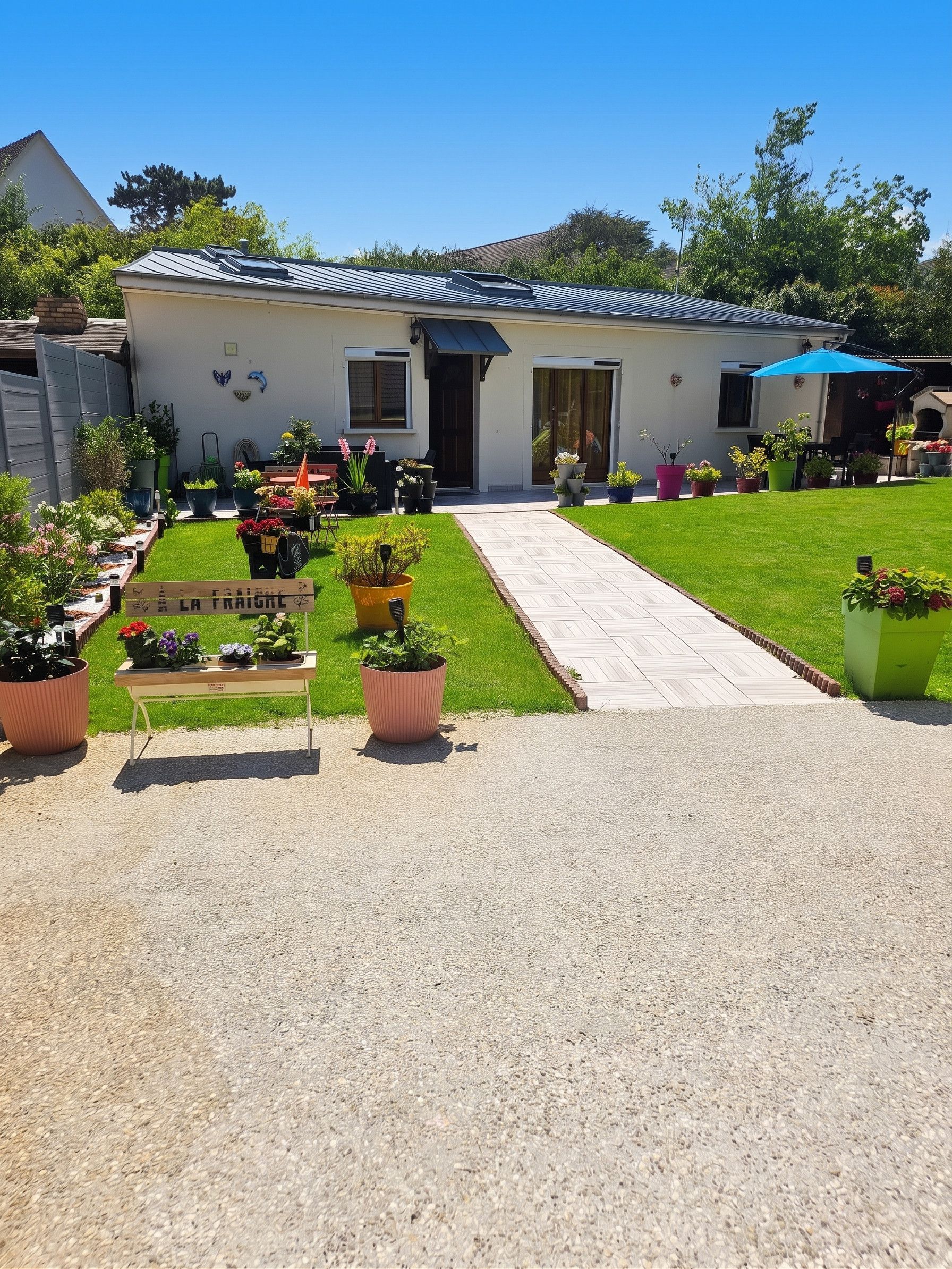 Una casa con un camino de entrada pavimentado que conduce a la puerta principal, rodeada de un césped cuidado y diversas macetas con plantas.