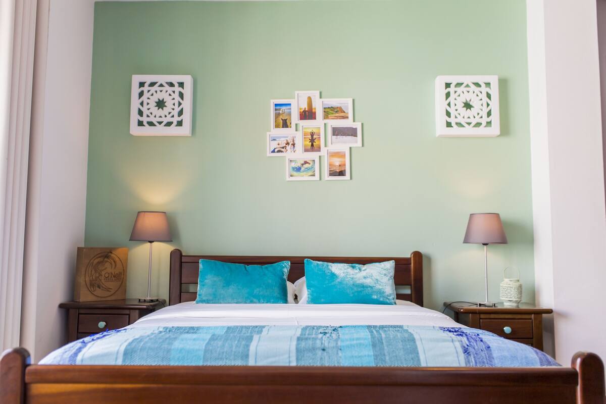 Ein ordentlich gemachtes Bett mit einer blau-weißen, gestreiften Tagesdecke, zwei passende Kissen und ein hölzernes Kopfteil. Es gibt zwei Nachttische mit Lampen, eine an der Wand montierte Leuchte sowie eine Reihe gerahmter Fotografien an der Wand.