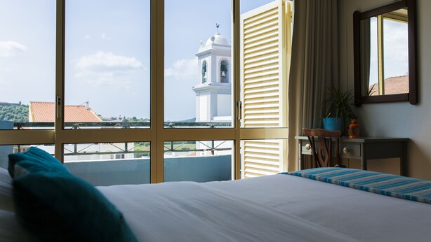 A hotel room with a large window, a bed, a bedside table, a chair, and a potted plant. - Atlantic Lodge (Aljezur)