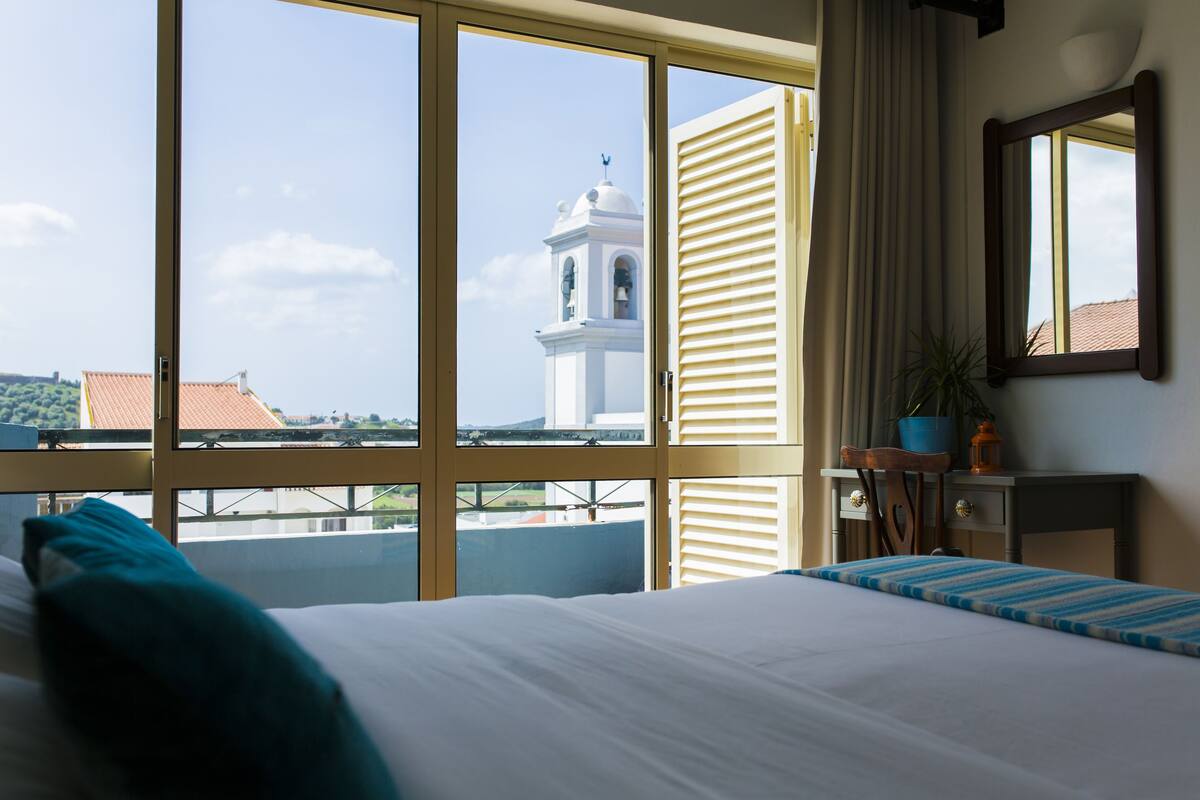 Ein Hotelzimmer mit großem Fenster, einem Bett, einem Nachttisch, einem Sessel und einer Zimmerpflanze.