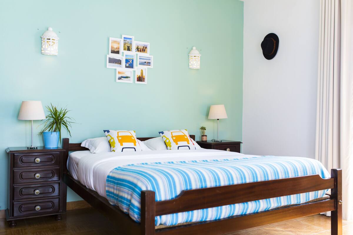 Ein ordentlich bezogenes Bett mit blau-weiß gestreifter Bettdecke, zwei Nachttische mit Lampen, eine Wand mit gerahmten Fotos und ein Blumentopf.