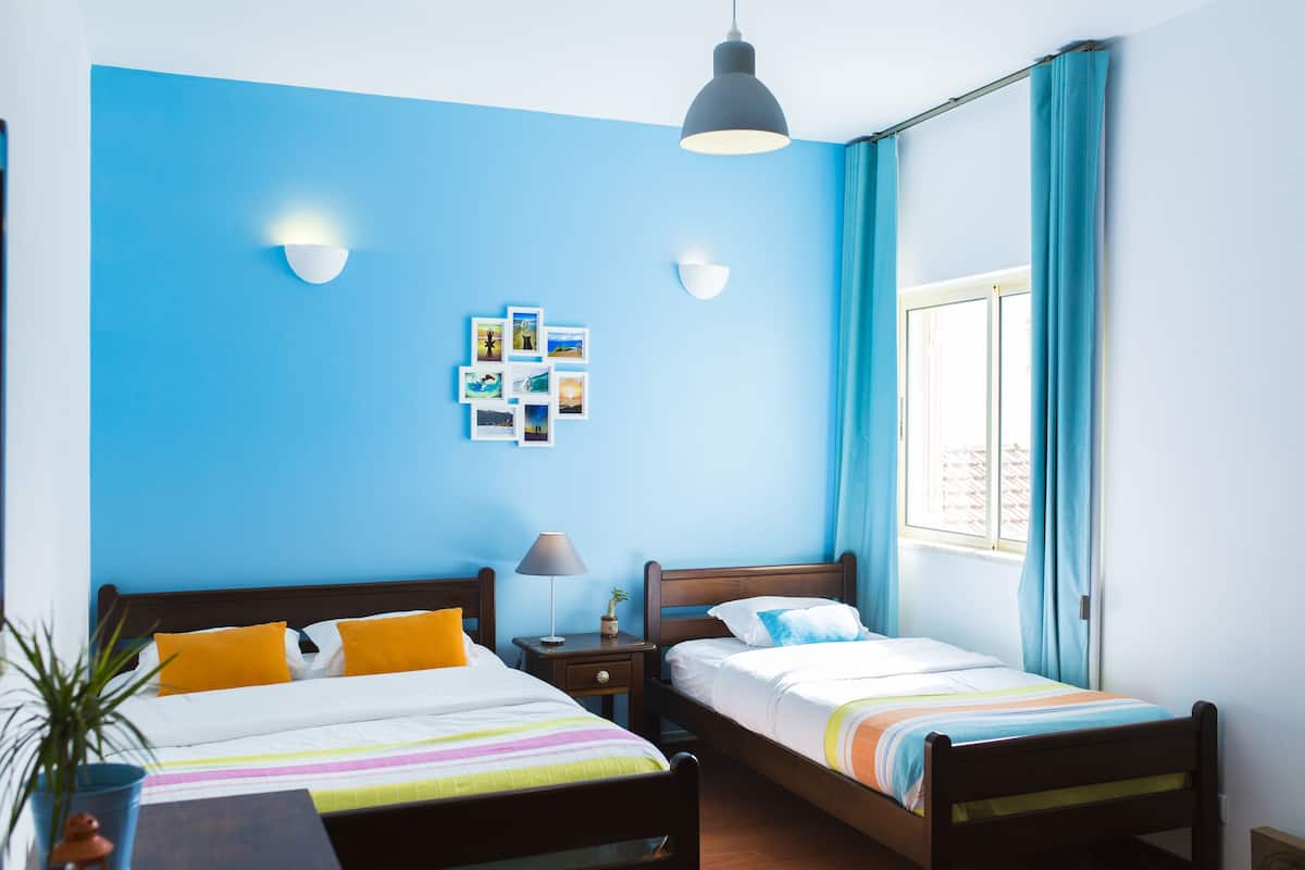 Ein Schlafzimmer mit zwei Betten, einem Nachttisch mit Lampe, einer Wandleuchte und einem Fenster mit Vorhängen.