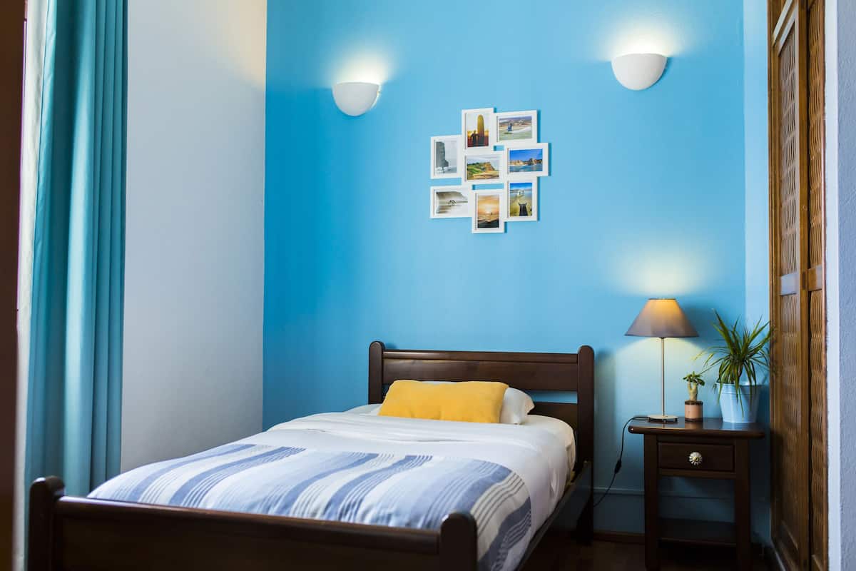 Ein ordentlich bezogenes Bett mit einer gestreiften Tagesdecke, einem gelben Kissen und einem hölzernen Kopfteil. Zwei Wandleuchten und ein kleiner Tisch mit Lampe und Topfpflanze sind vorhanden.
