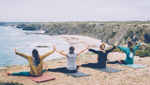 Three people doing yoga on mats by a beach with a rocky cliff in the background. - Atlantic Lodge (Aljezur)