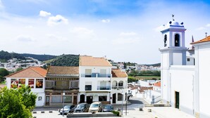 A white building with a bell tower, a tiled roof, and a view of a town in the background. - Atlantic Lodge (Aljezur)