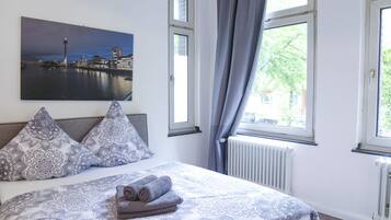 Ein Schlafzimmer mit Bett, gemusterten Kissen, einem Stadtbildgemälde und einem Fenster mit Vorhängen.