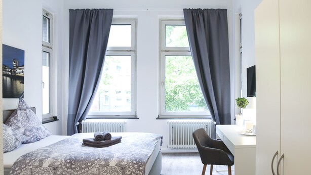 Un dormitorio con cama, escritorio y silla. Hay dos ventanas con cortinas, un radiador y una planta sobre el escritorio.