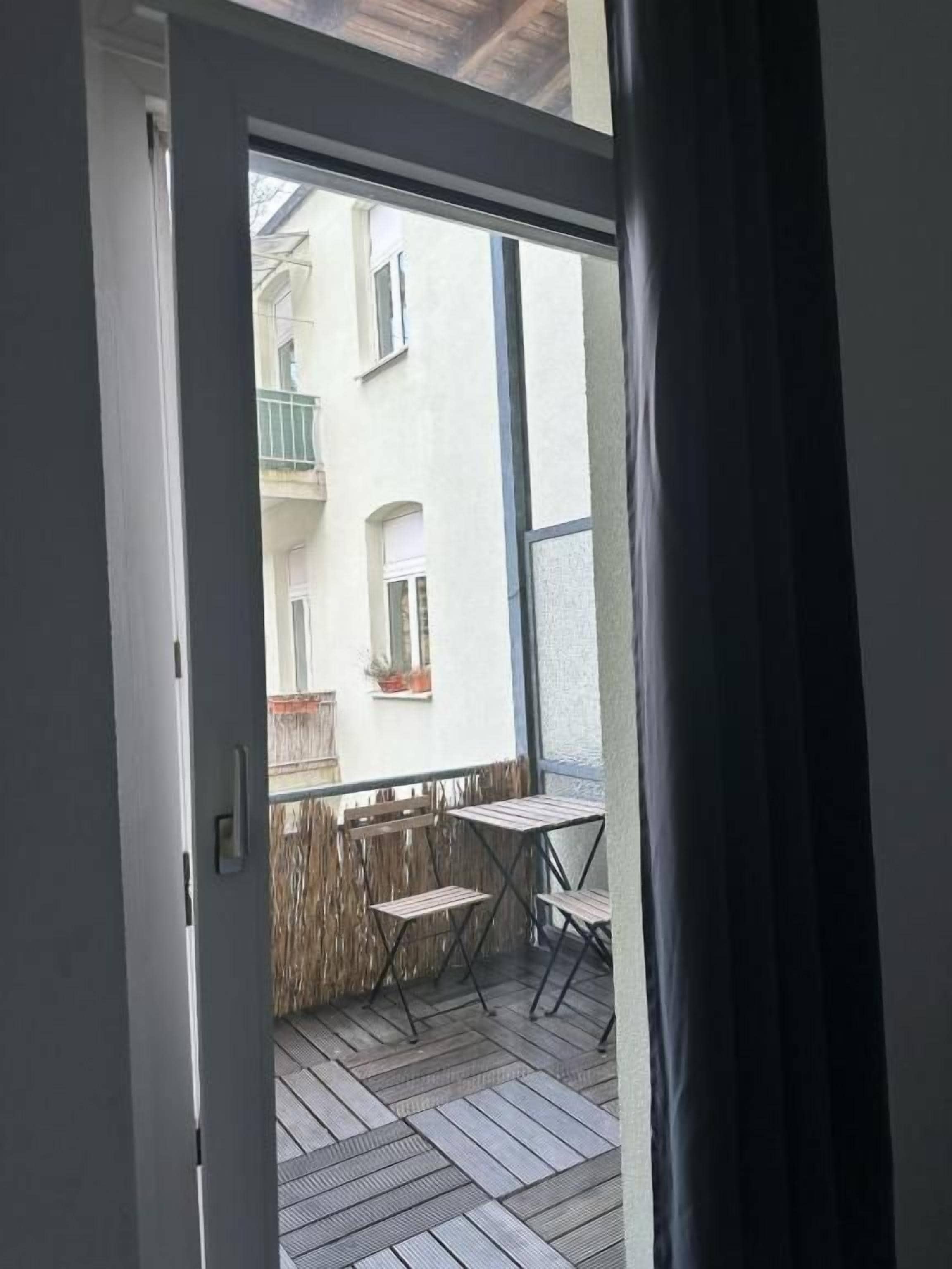 An open door to a balcony with a view of a building, a table, and two chairs.