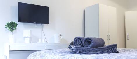 Quarto de hotel com cama, televisão de tela plana na parede, guarda-roupa, escrivaninha com abajur e uma pequena planta em vaso.