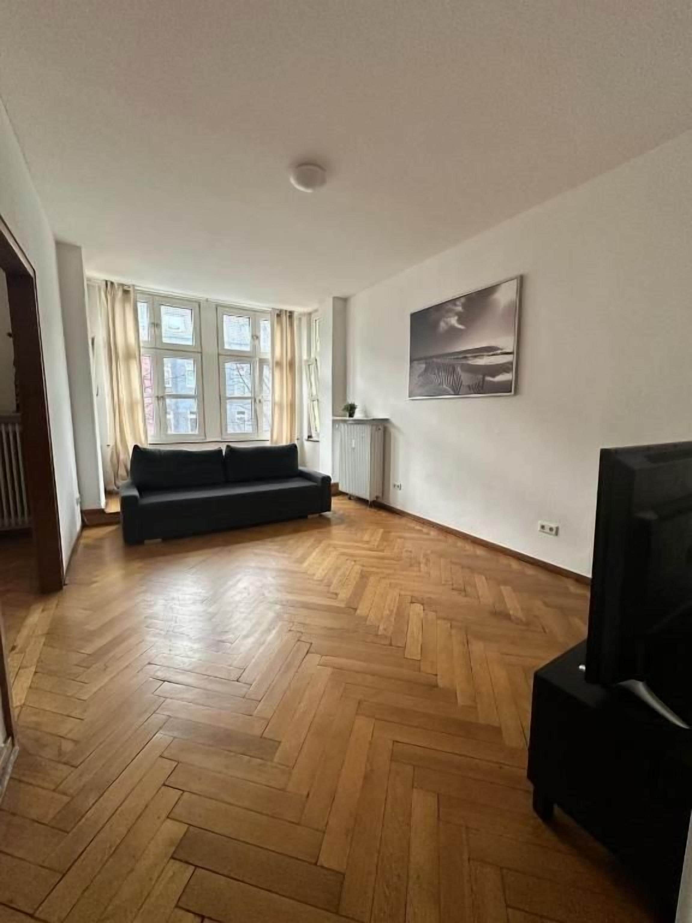 A spacious living room with a herringbone floor, a black sofa, a television, and a window with curtains.