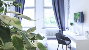Una habitación luminosa con un ventanal grande, un radiador, una silla negra y un televisor en la pared.