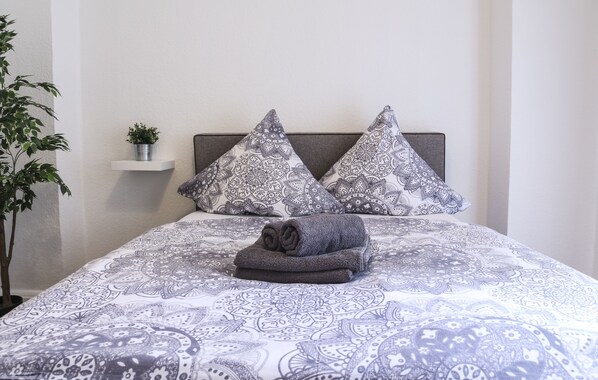 Una cama bien hecha con ropa de cama estampada, dos almohadas y una toalla enrollada en una pared blanca con un estante que sostiene una planta.