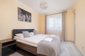 Un dormitorio con cama, mesita de noche, un cuadro y ventana con cortinas.