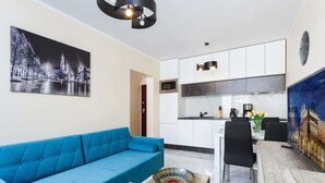 A modern living room with a blue sofa, a coffee table, and a dining area with chairs. There is a kitchen with white cabinets and a painting of a cityscape on the wall.