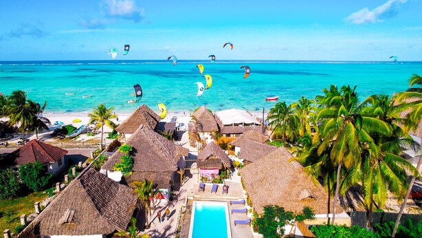 Un resort en la playa con cabañas de techo de paja, piscina y vista al océano con surfistas de kitesurf.