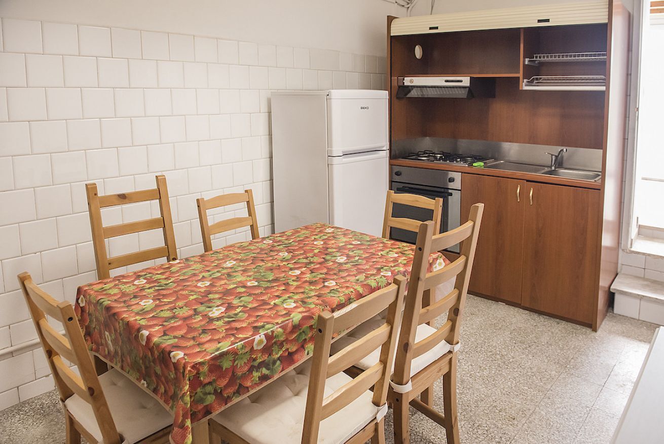 Een kleine keuken met een koelkast, fornuis en wastafel. Er is een eettafel met stoelen.
