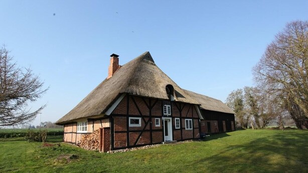 Une maison traditionnelle à colombages, avec un toit de chaume, une cheminée et une porte surmontée d’une petite fenêtre.