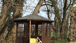 Une petite cabane en bois avec un banc à l’intérieur, entourée d’arbres et d’un mur de pierres.
