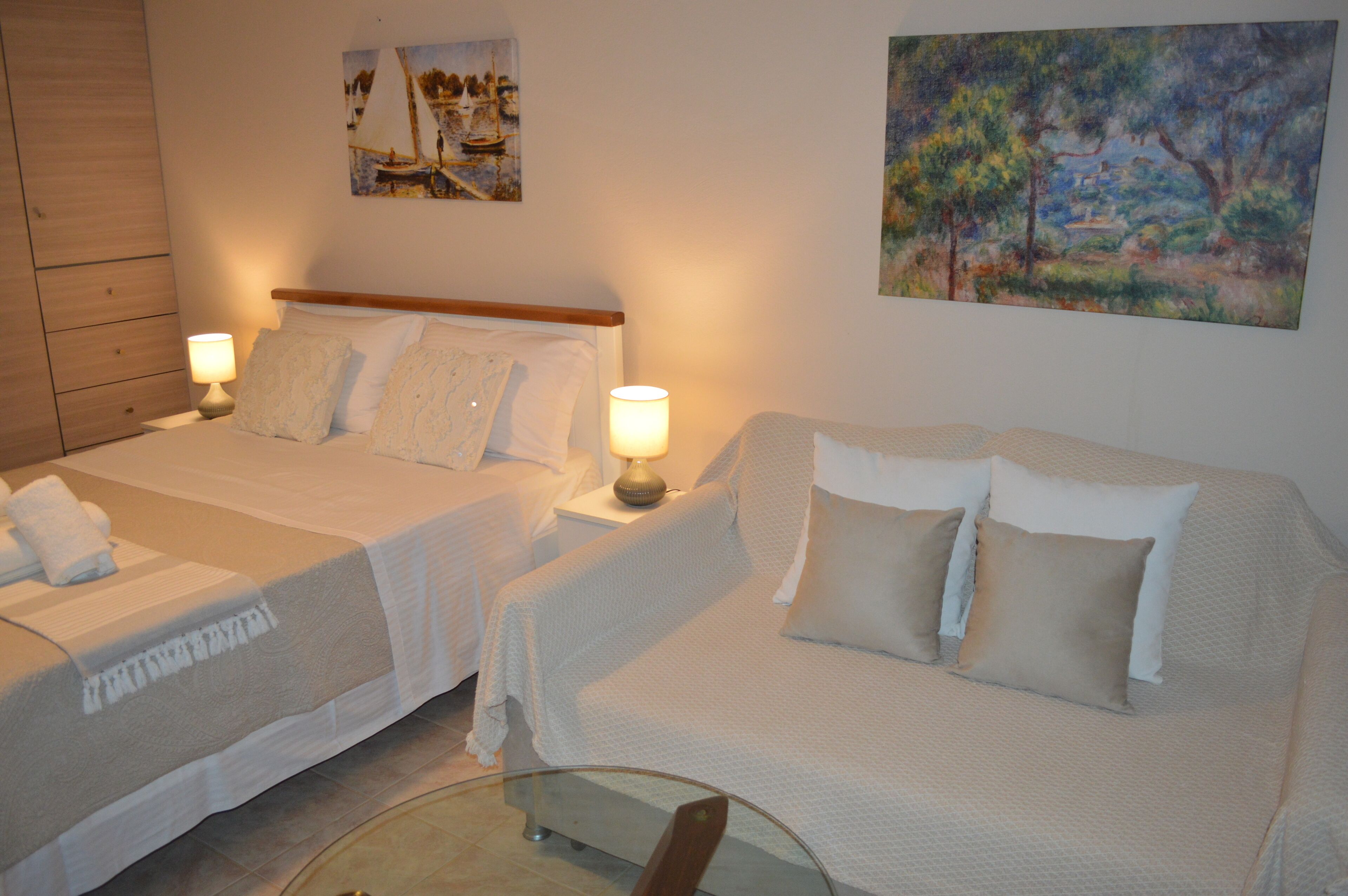 A hotel room with two beds, a sofa, and a glass table. There are paintings on the walls and a lamp on each bedside table.