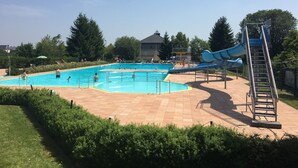Une piscine avec un toboggan et une échelle, entourée de verdure, avec un bâtiment visible à l’arrière-plan.