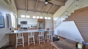 A modern kitchen with a central island, white bar stools, a wooden dining table, and a staircase leading to an upper level.