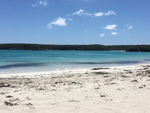Een strand met helder turquoise water, wit zand en een veraf gelegen bebost gebied.