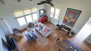 A spacious living room with a high ceiling, a ceiling fan, a large window, a fireplace, and a colorful quilt hanging on the wall.