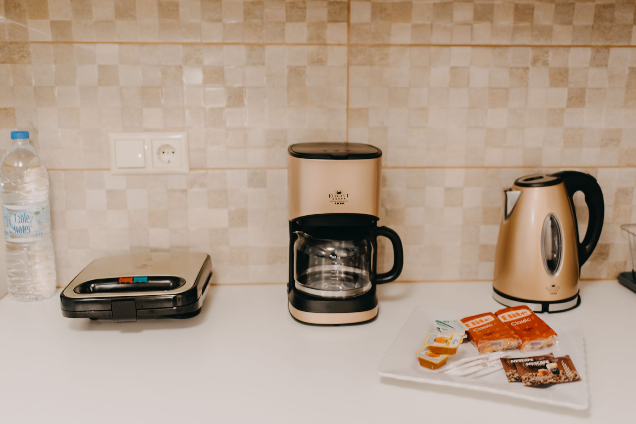 Un plan de travail de cuisine avec une machine à café, une bouilloire, une balance et une bouteille d’eau.
