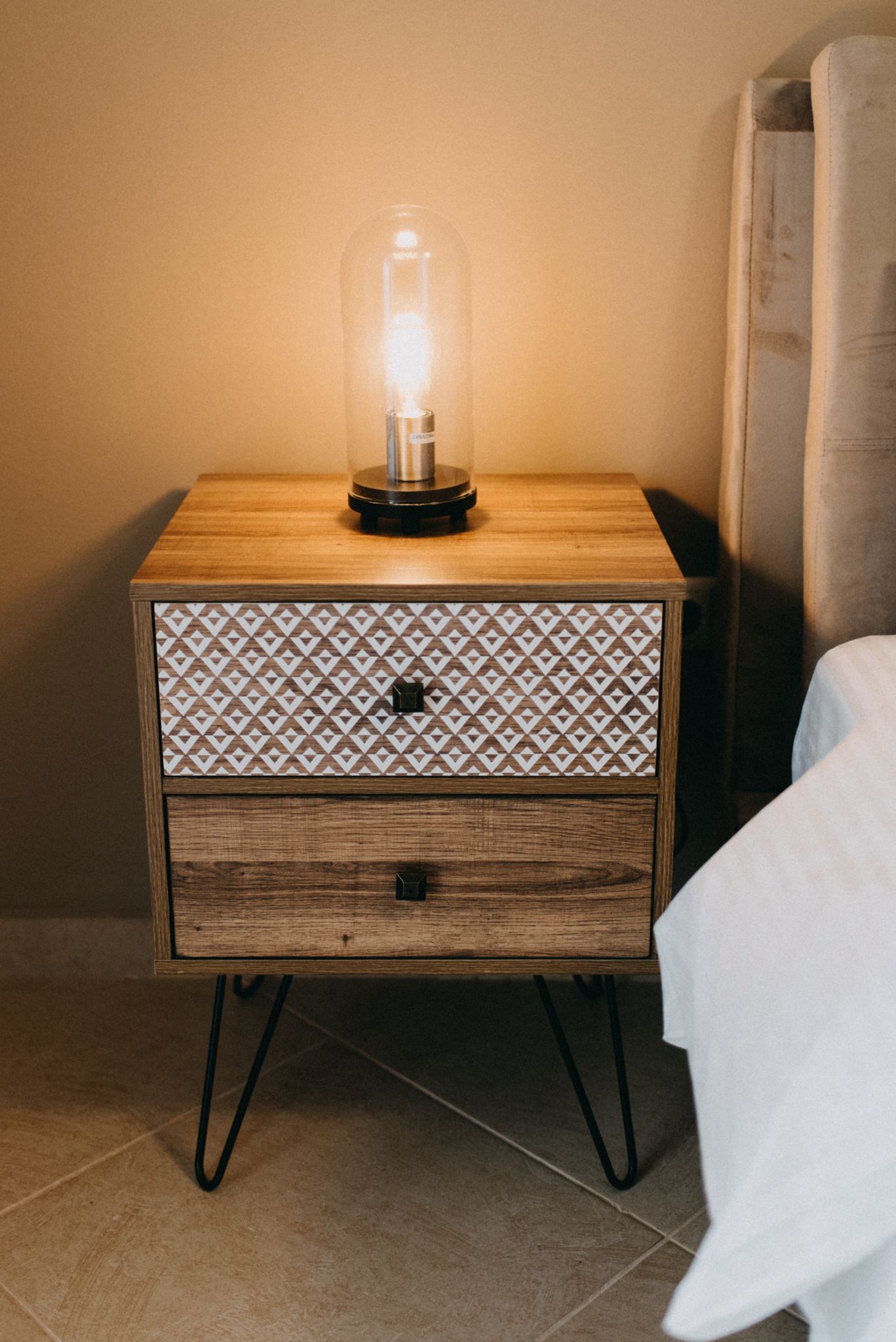 A wooden bedside table with a glass bulb lamp, a patterned drawer, and metal legs.