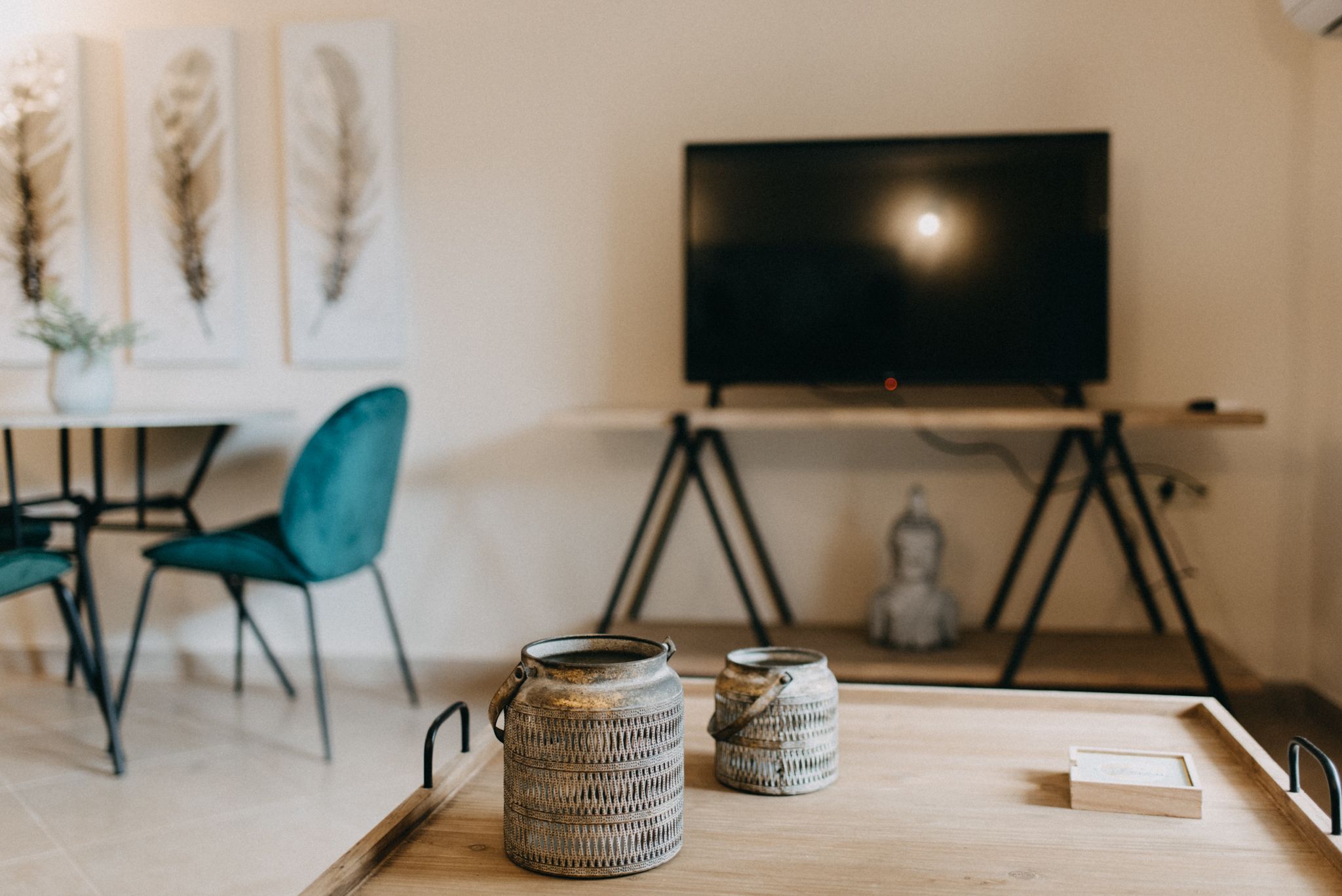 A modern living room with a wooden coffee table, two ceramic vases, a teal chair, a black TV on a tripod stand, and a framed artwork on the wall.