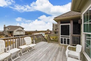A wooden deck with white chairs, a glass door, and a view of the beach.