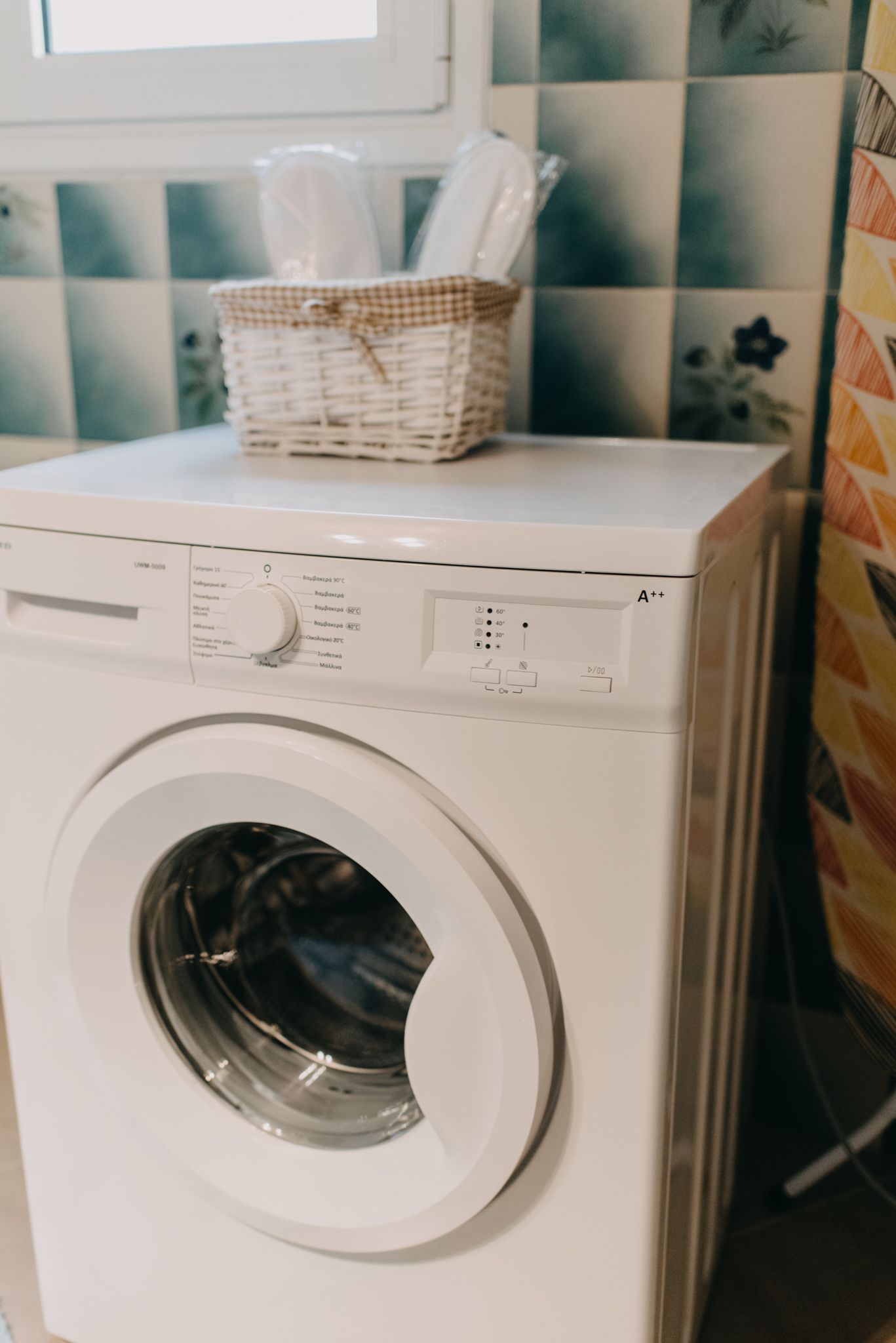 A front-loading washing machine with a control panel, a basket on top, and a tiled backsplash in the background.