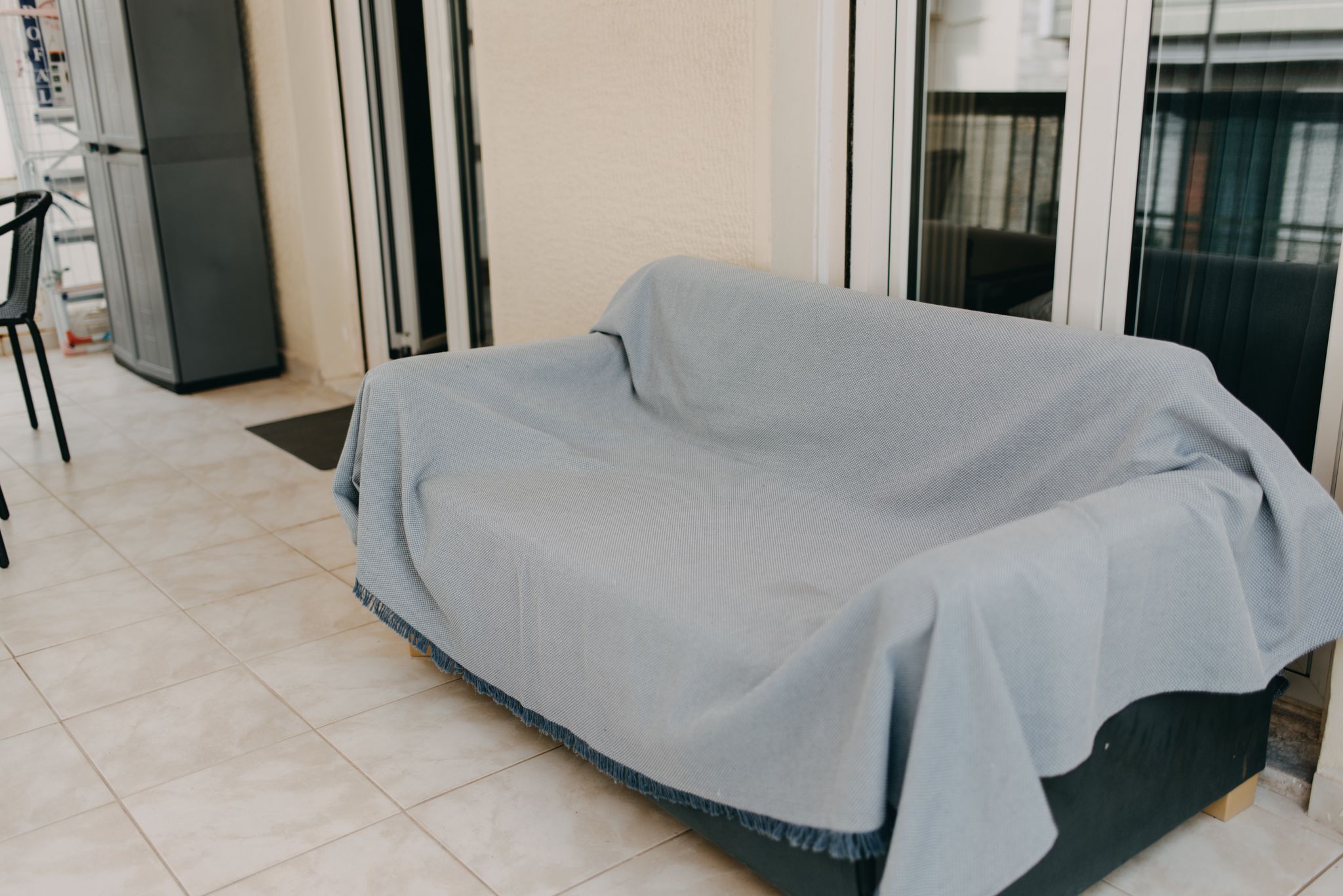Un sofá cubierto en una habitación con suelo de baldosas, una puerta y una silla.
