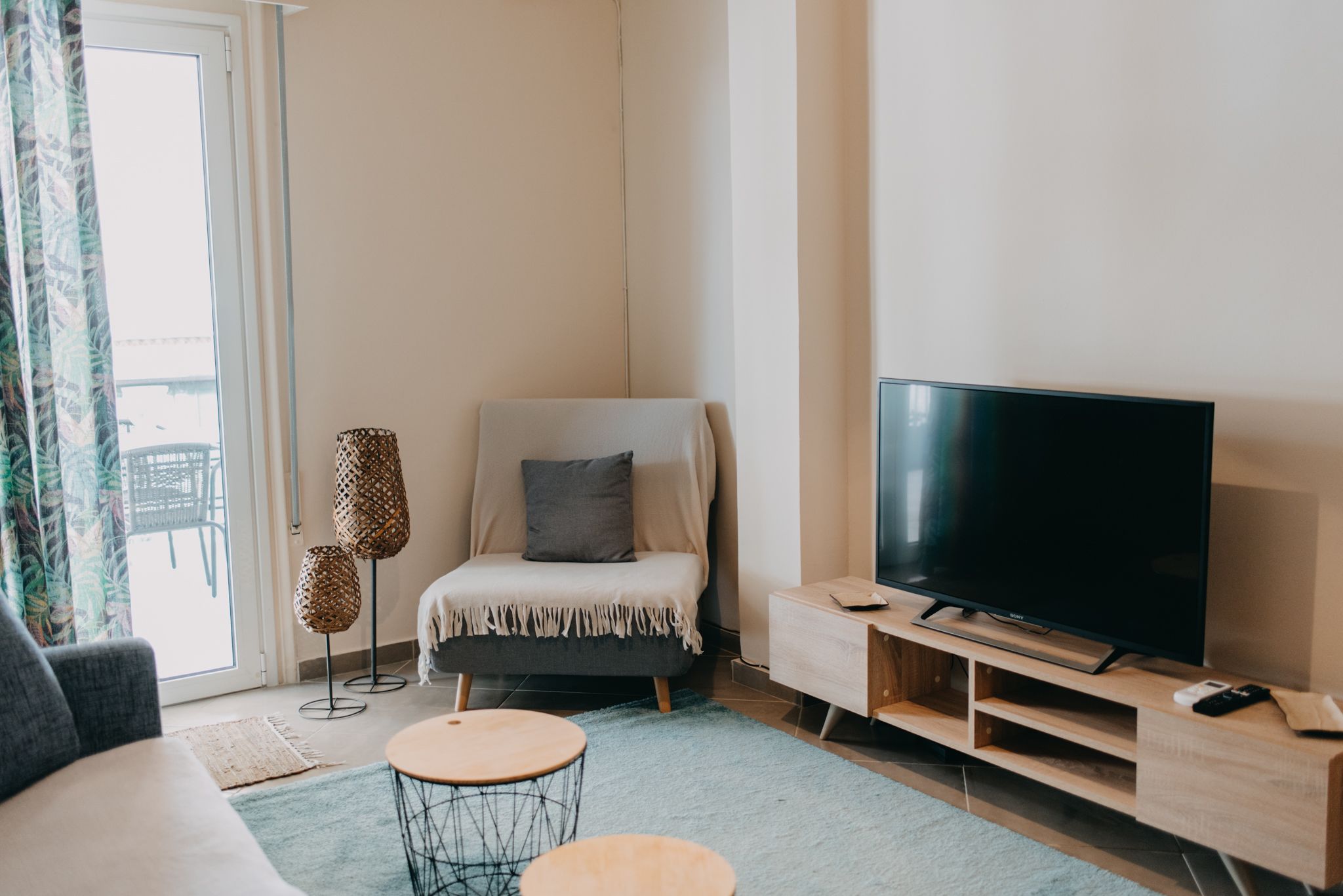 A modern living room with a sofa, a small round table, a TV on a wooden stand, and a balcony with a chair and a patterned floor mat.