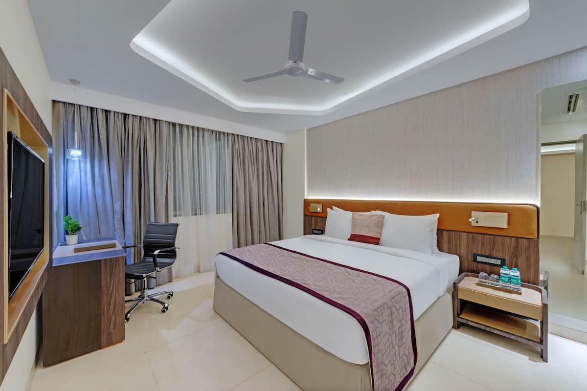 A hotel room with a bed, desk, chair, TV, and a nightstand with bottles.