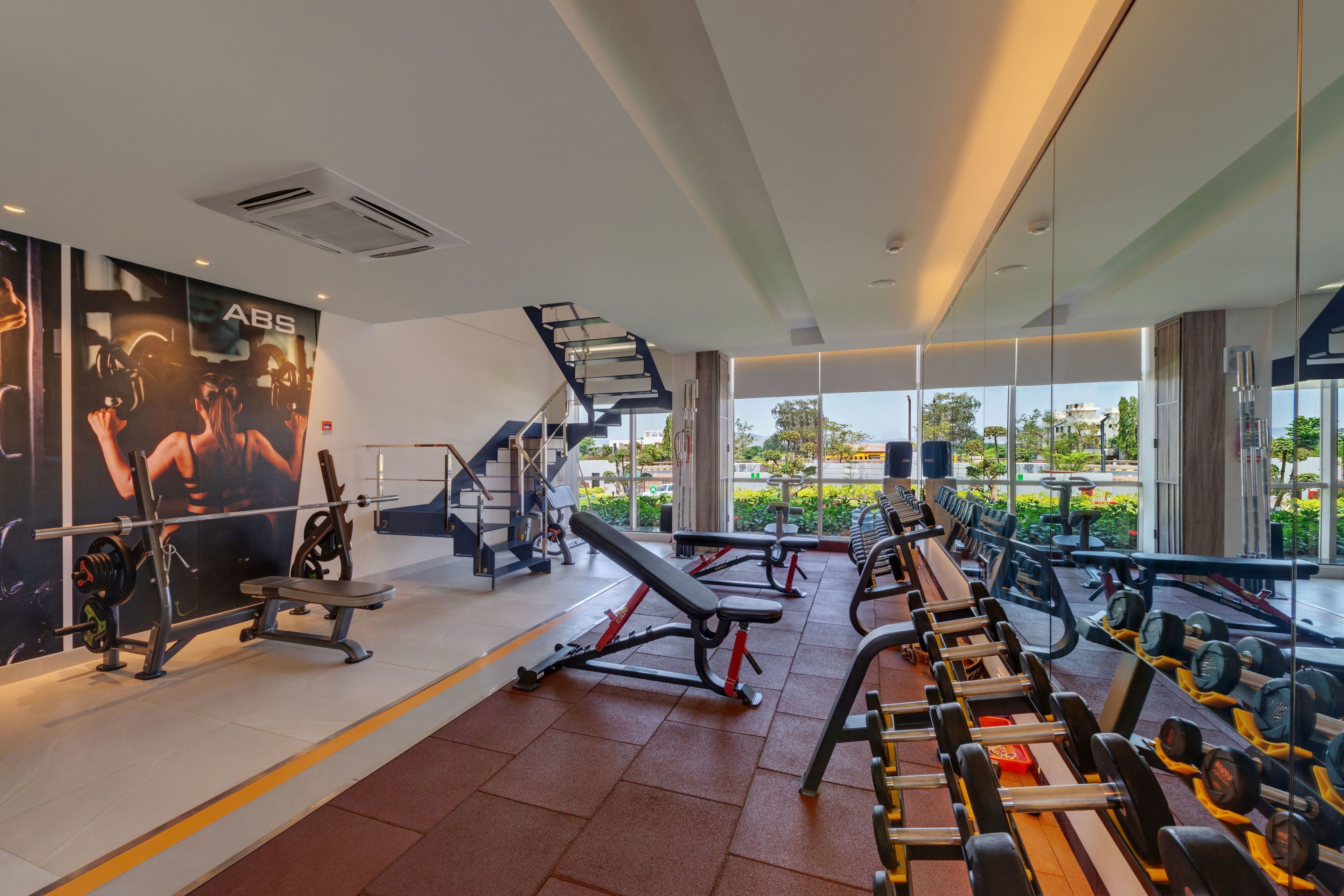 A modern gym with various exercise equipment, a staircase, and large windows offering a view of the outdoors.