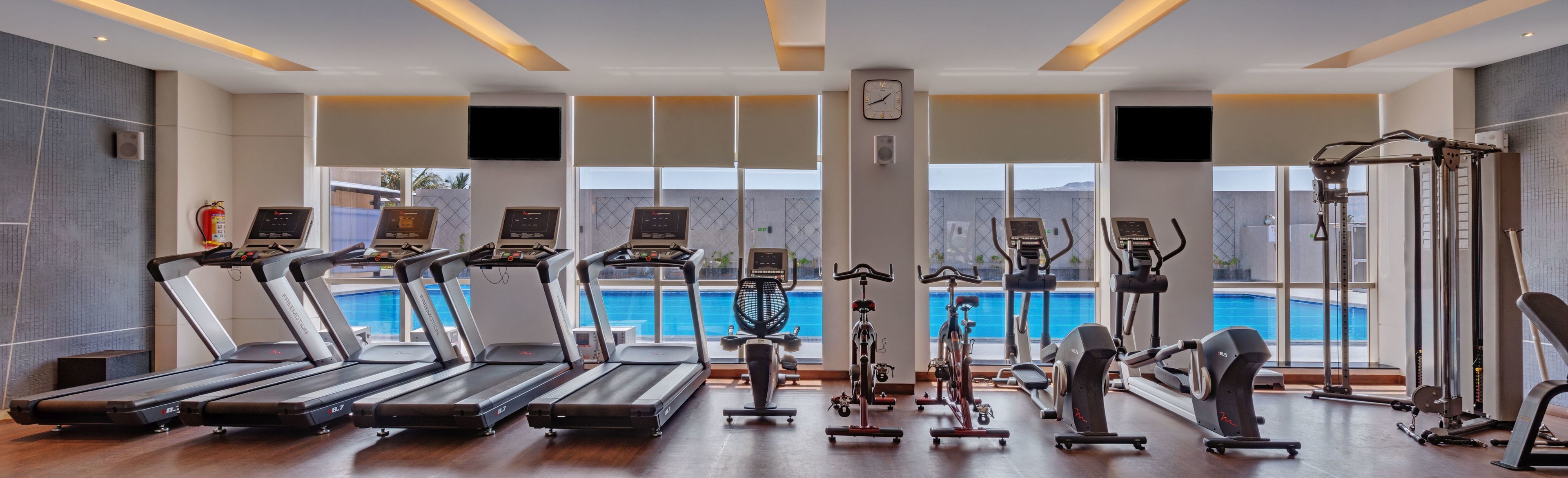 A gym with treadmills, stationary bikes, and weight machines, overlooking a swimming pool.