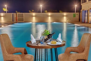 A poolside table with a bottle, glasses, and a slice of cake, surrounded by two chairs.