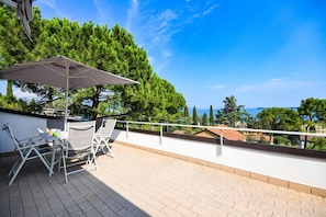 A terrace with a table and chairs, a large umbrella, and a view of the sea and trees. - Cupido (Gardone Riviera)